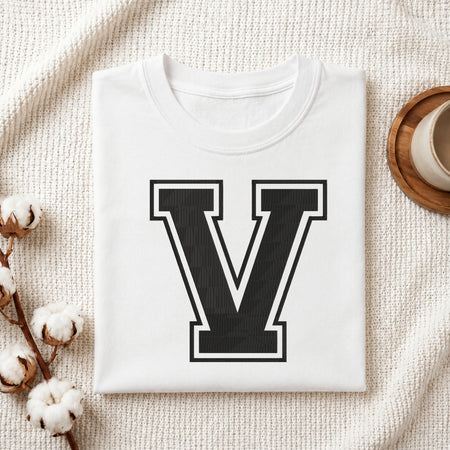 A person carrying a white canvas tote bag featuring a large embroidered varsity block letter 'V' in black with a white outline, set in a bright room with green house plants.