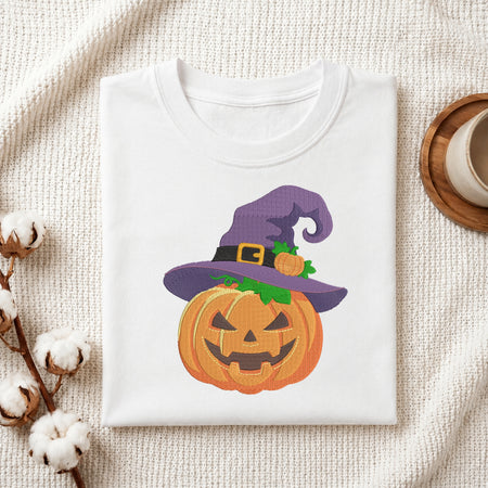 A folded white t-shirt with a Halloween graphic of a jack-o'-lantern wearing a purple witch hat, styled on a textured cream fabric with cotton stems and a coffee cup.