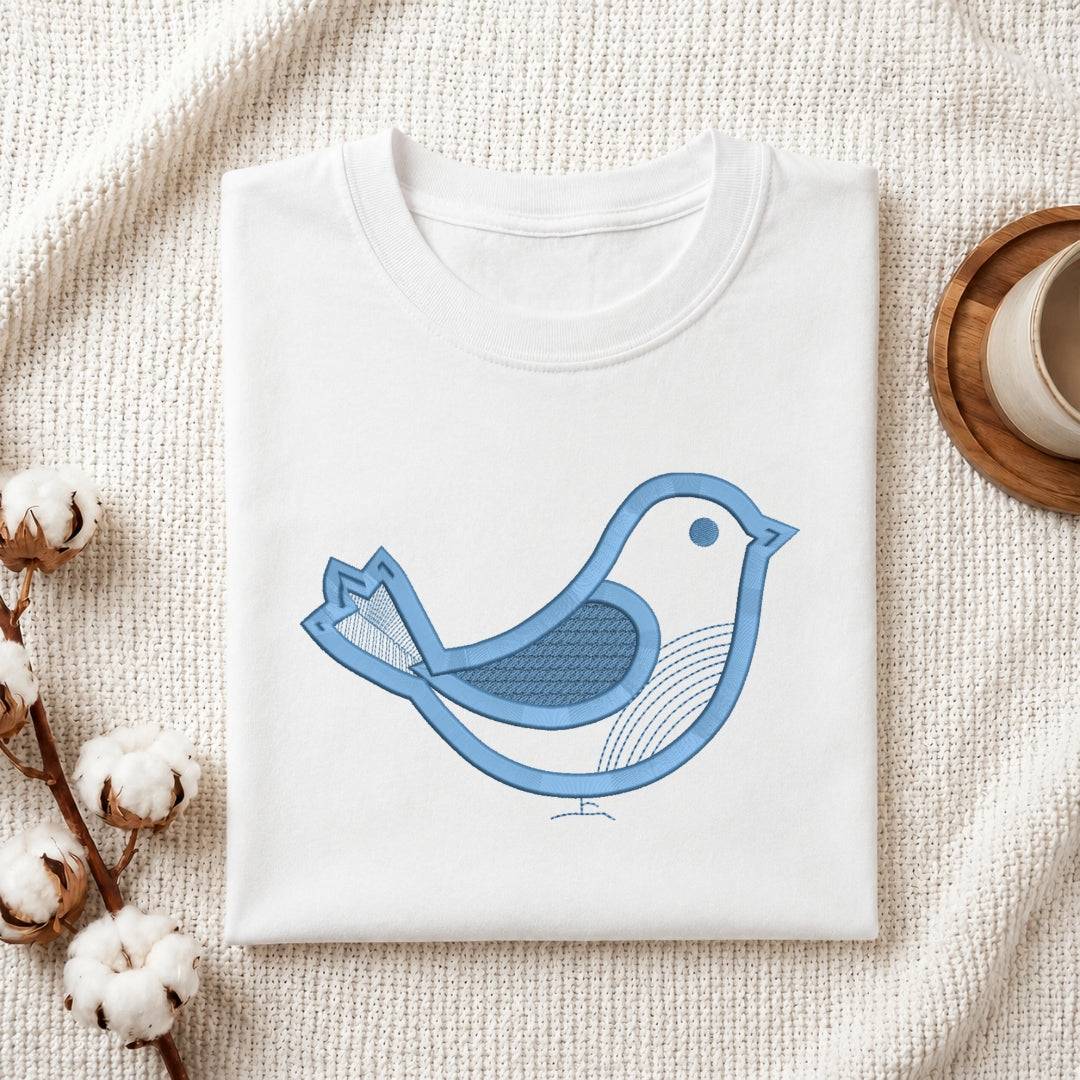 A folded white crewneck t-shirt featuring a blue stylized bird graphic on the front, styled on a textured cream knit blanket with cotton bolls and a wooden cup in the background.