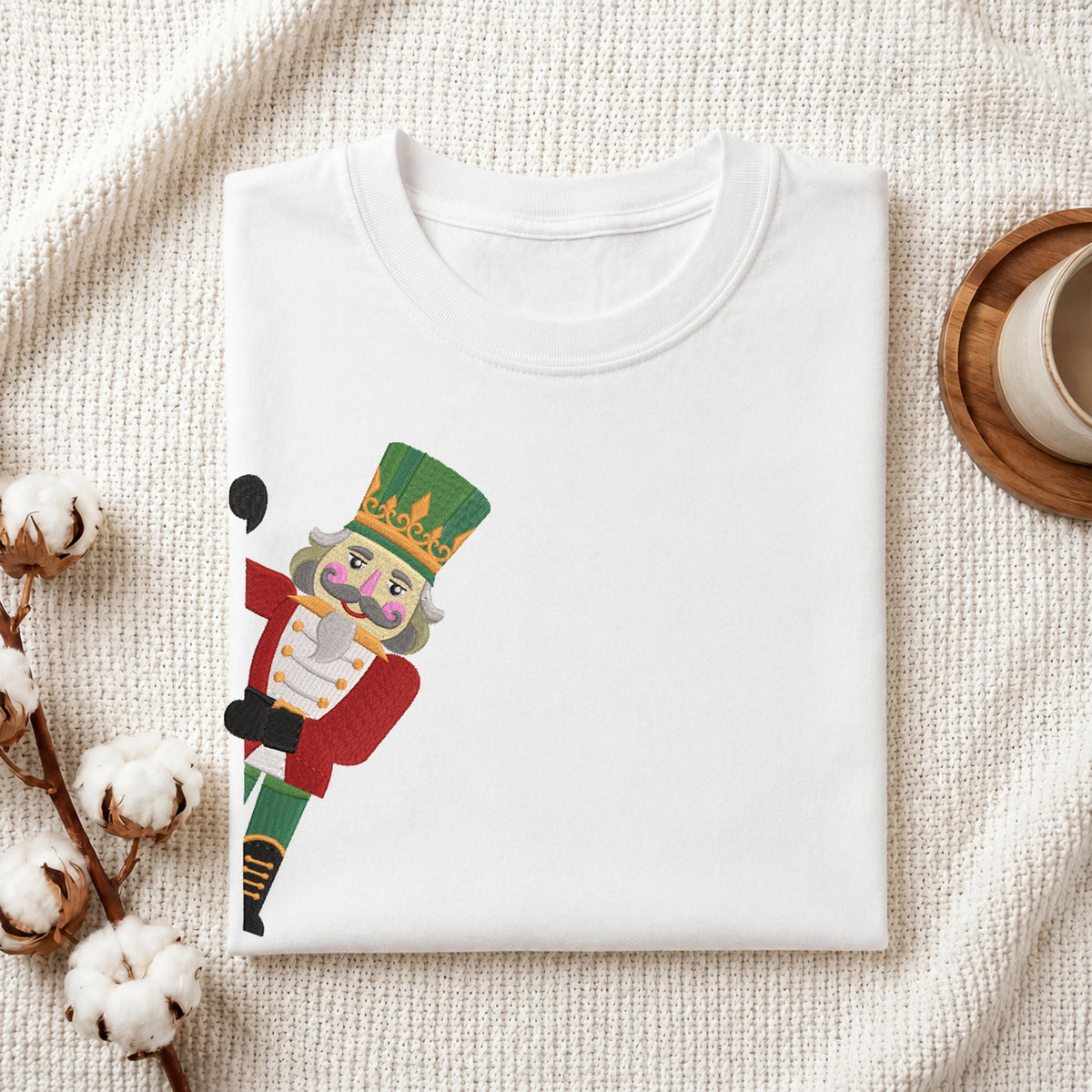 A neatly folded white t-shirt featuring the embroidered peeking nutcracker soldier graphic, displayed on a textured white knit background with cotton stems and a wooden cup.