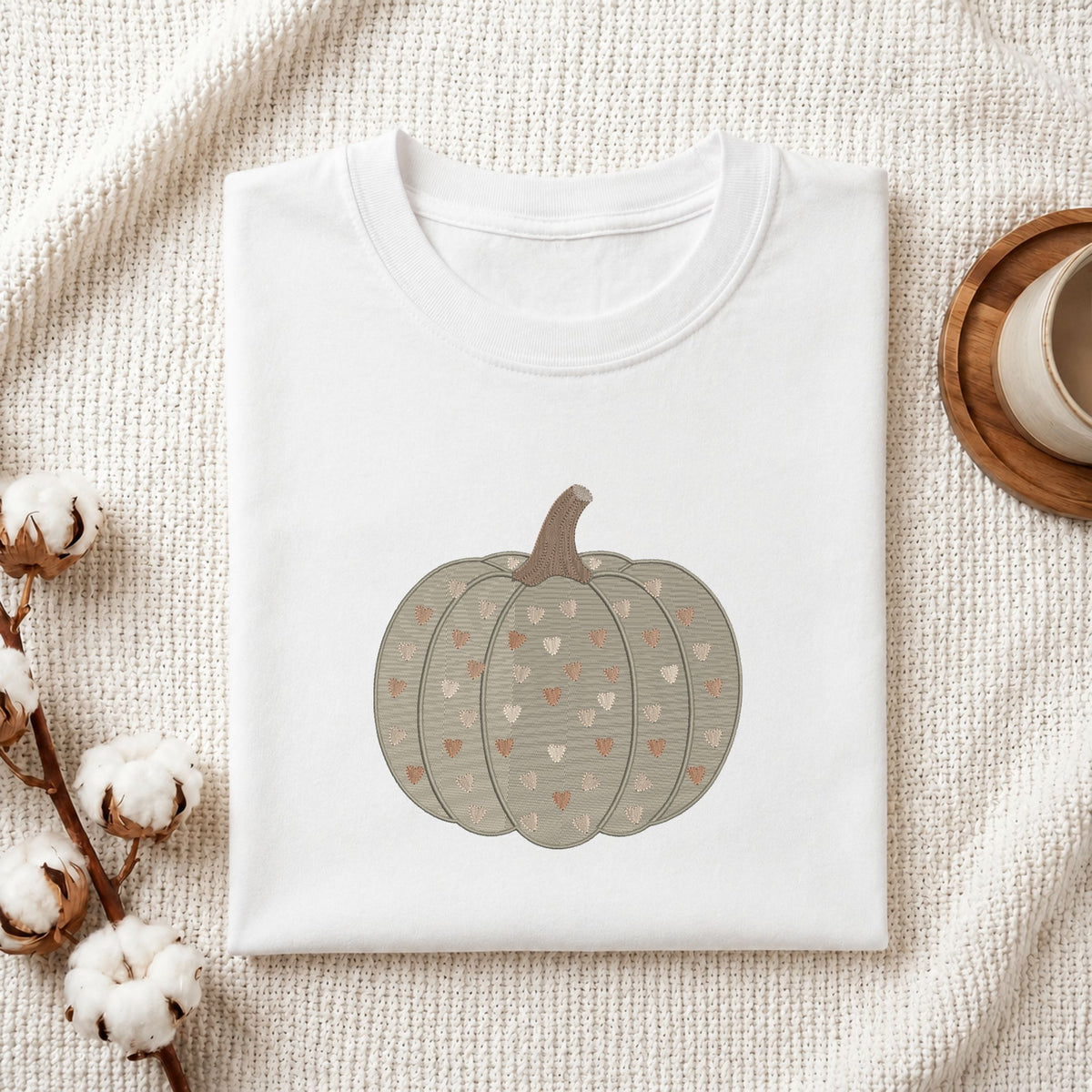 A folded white crewneck t-shirt featuring a printed graphic of a grayish-green pumpkin with small hearts, styled on a textured cream fabric with cotton bolls and a wooden cup.