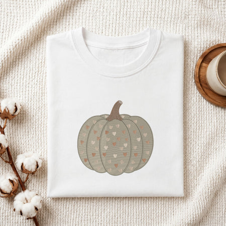 A folded white crewneck t-shirt featuring a printed graphic of a grayish-green pumpkin with small hearts, styled on a textured cream fabric with cotton bolls and a wooden cup.