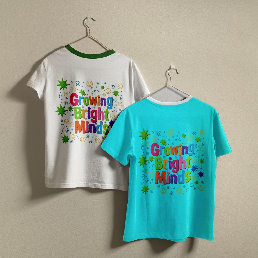 Two children's short-sleeve T-shirts hanging on hangers against a plain beige wall. One shirt is white with a green collar and a colorful back graphic reading 'Growing Bright Minds'; the other is bright turquoise with a white collar and a matching colorful front graphic.