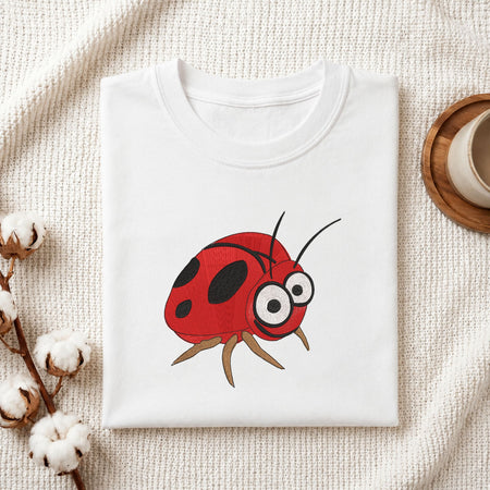 Folded white cotton t-shirt mockup featuring a cute ladybug machine embroidery design, styled with cotton stems and a wooden cup