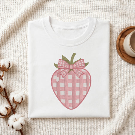 A folded white crewneck T-shirt displayed on a textured cream fabric background, featuring a pink and white gingham strawberry graphic with a matching bow and green leaves.