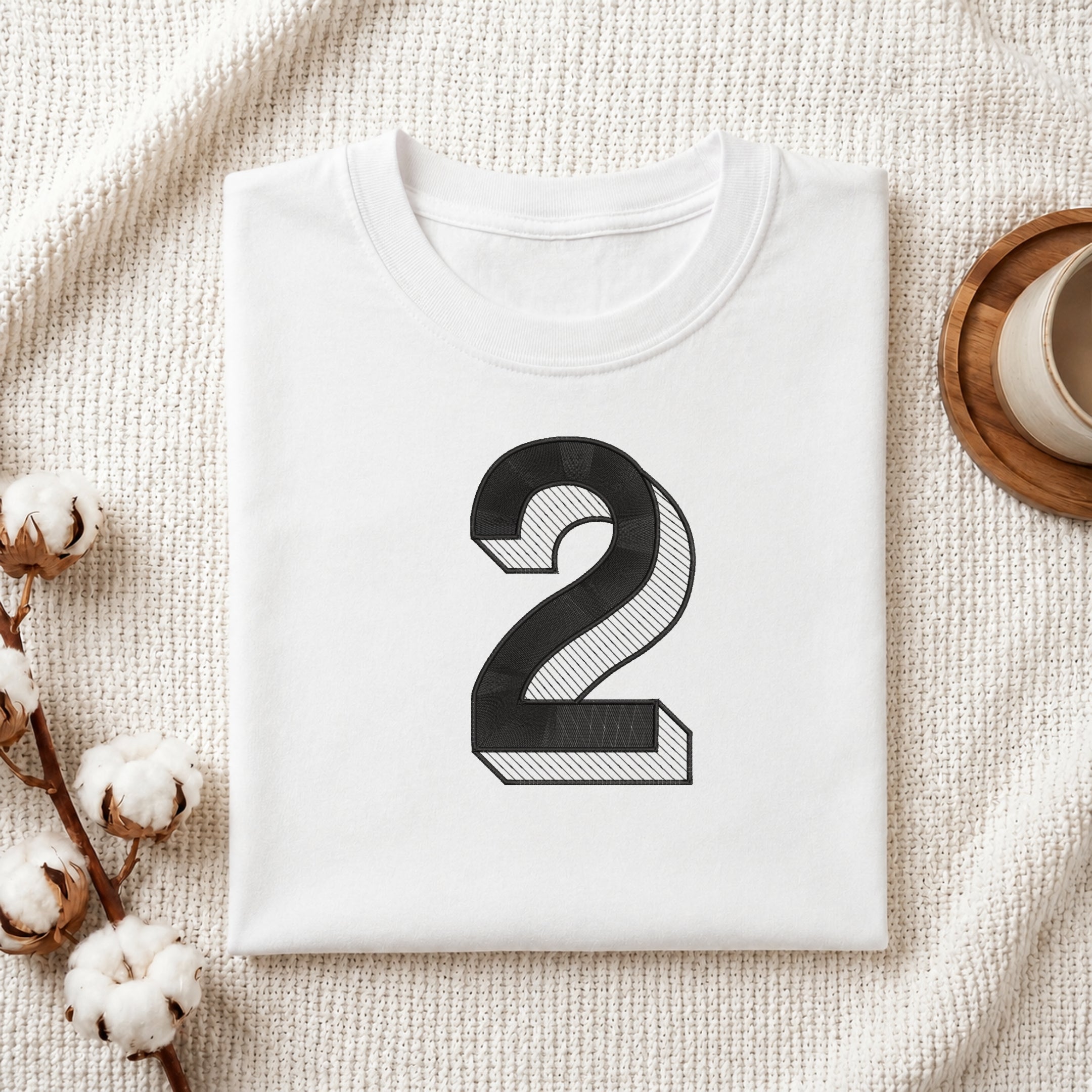 A folded white T-shirt with a large black number 2 printed on the front, styled on a textured cream fabric with cotton stems and a small cup in the corners.