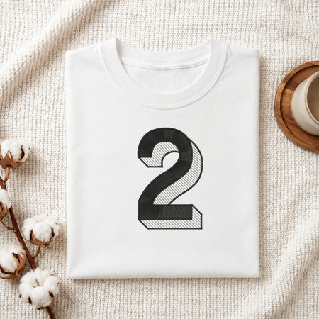 A folded white T-shirt with a large black number 2 printed on the front, styled on a textured cream fabric with cotton stems and a small cup in the corners.