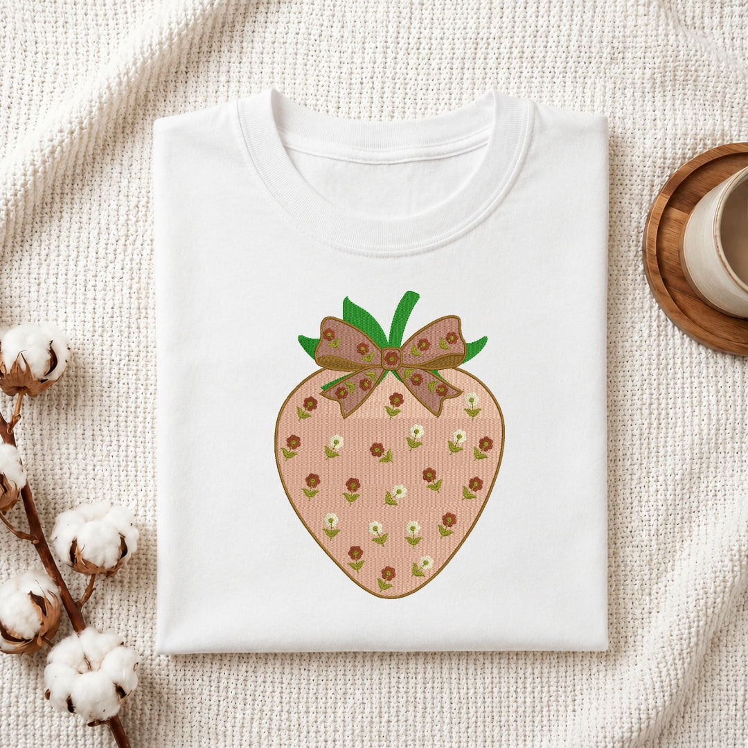 A folded white crewneck t-shirt featuring a graphic of a strawberry with a brown patterned ribbon bow, styled on a textured cream fabric with cotton stems and a wooden cup.
