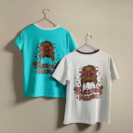Two children's graphic T-shirts hanging on hangers against a plain beige wall. One shirt is turquoise blue and the other is white, both featuring a graphic of a woman with a brown hair bun and pink bow, with the text 'Blessed Mama' in pink and brown.
