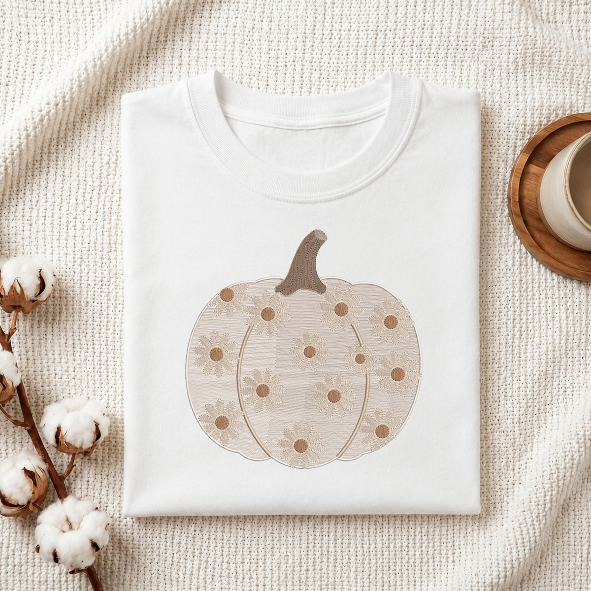 A folded white crewneck sweatshirt or t-shirt with a graphic of a beige pumpkin decorated with daisy-like flowers, styled on a textured cream fabric with cotton stems and a wooden cup in the background.