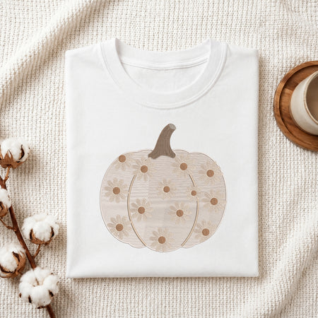 A folded white crewneck sweatshirt or t-shirt with a graphic of a beige pumpkin decorated with daisy-like flowers, styled on a textured cream fabric with cotton stems and a wooden cup in the background.