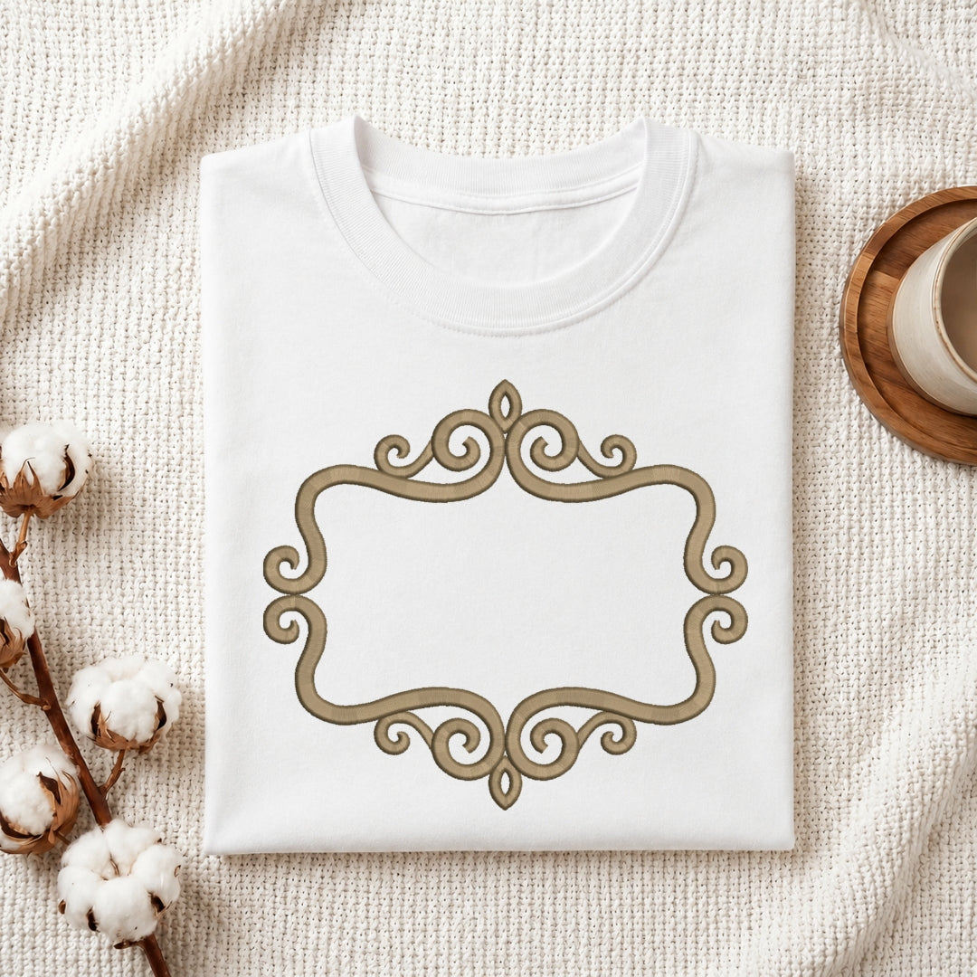 A folded white crewneck t-shirt with a decorative gold scrollwork frame on the front, styled on a textured cream blanket with cotton bolls and a wooden cup in the background.