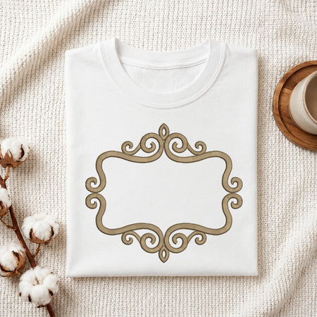 A folded white crewneck t-shirt with a decorative gold scrollwork frame on the front, styled on a textured cream blanket with cotton bolls and a wooden cup in the background.