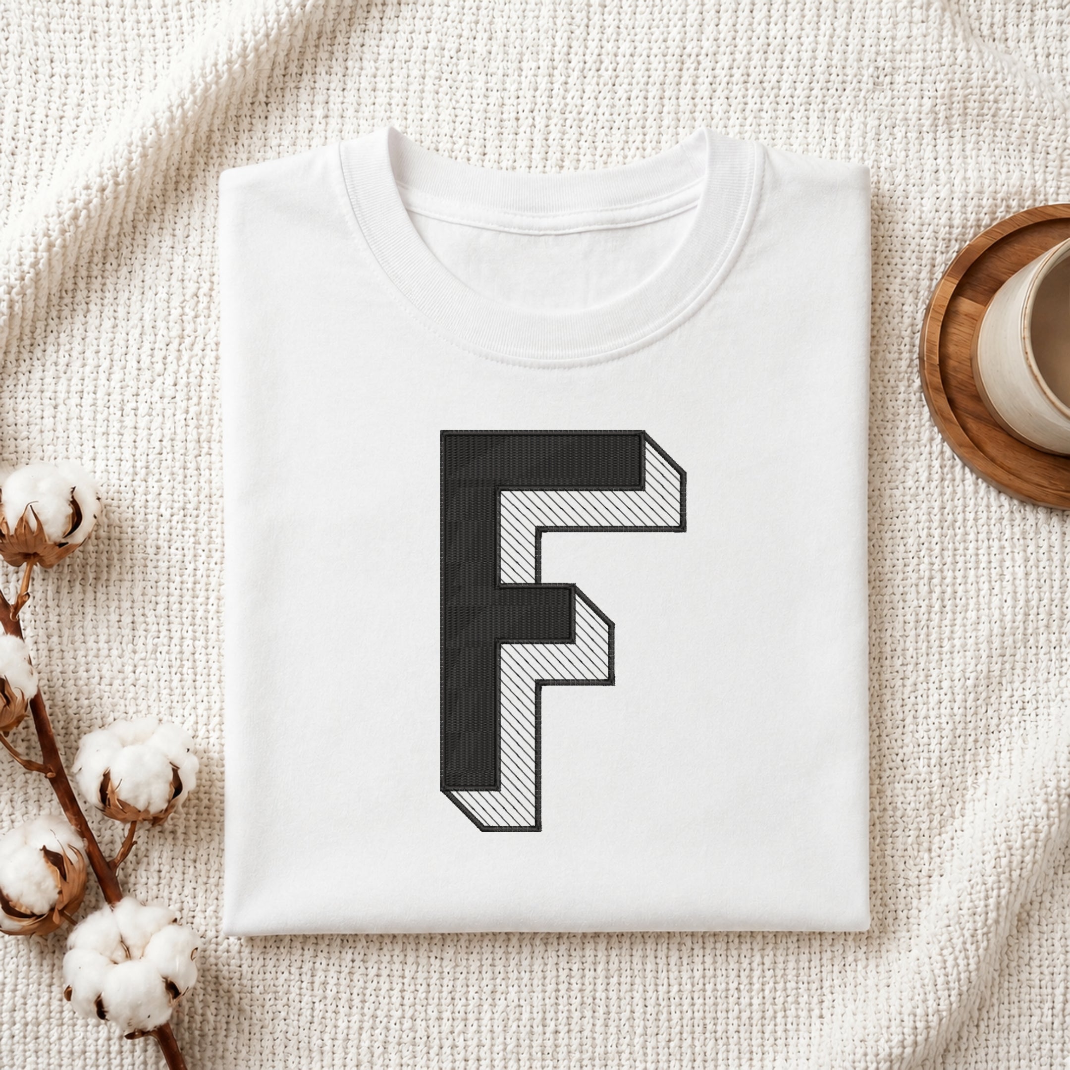 A folded white t-shirt with a large black block letter 'F' printed on the front, styled on a textured cream knit blanket with cotton stems and a cup of coffee in the background.
