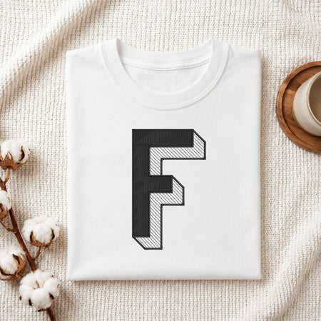 A folded white t-shirt with a large black block letter 'F' printed on the front, styled on a textured cream knit blanket with cotton stems and a cup of coffee in the background.