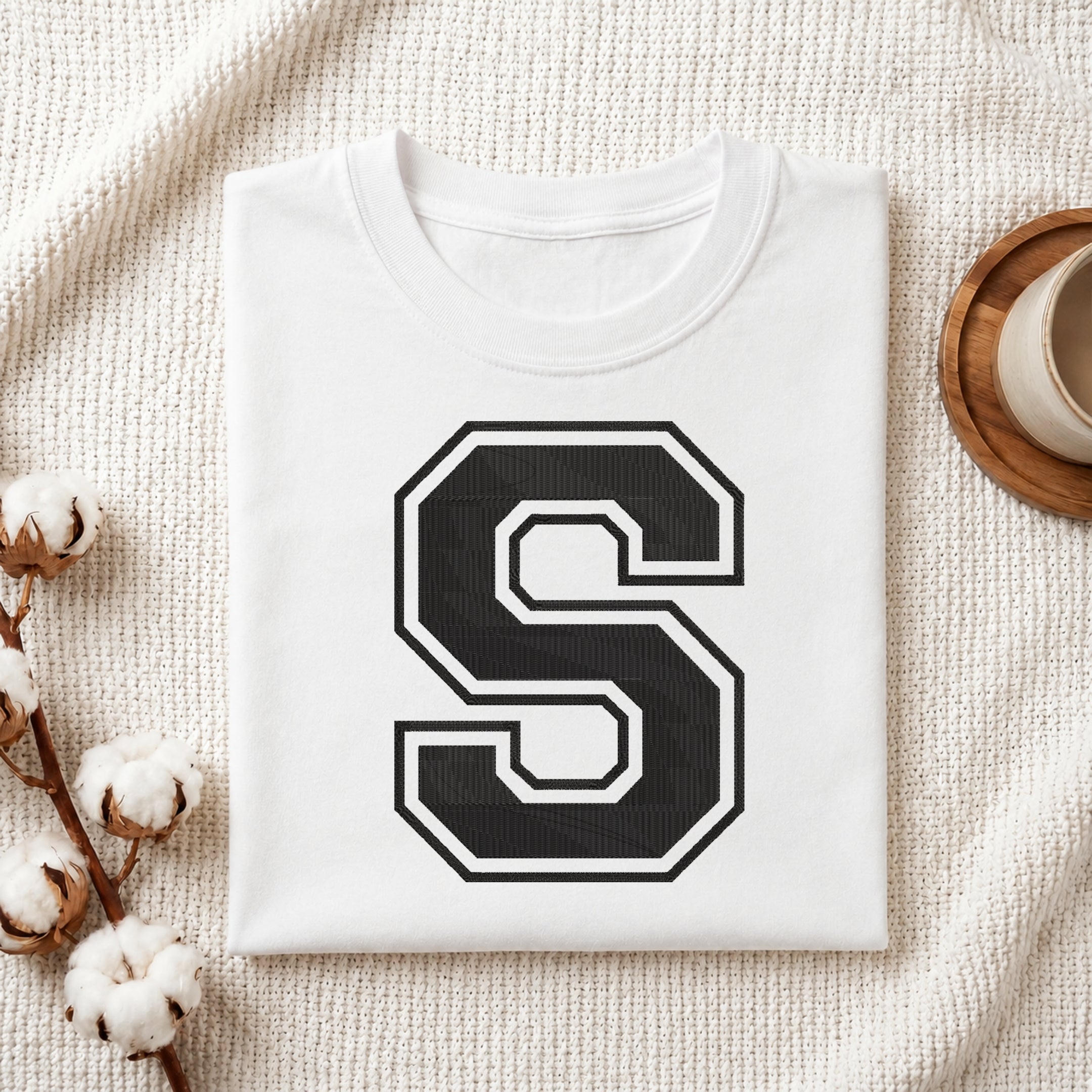 Flat-lay mockup showing a folded white t-shirt featuring a large collegiate-style embroidered black letter 'S' with a white border outline, set against a cozy white knitted blanket background.