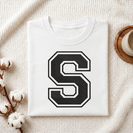 Flat-lay mockup showing a folded white t-shirt featuring a large collegiate-style embroidered black letter 'S' with a white border outline, set against a cozy white knitted blanket background.