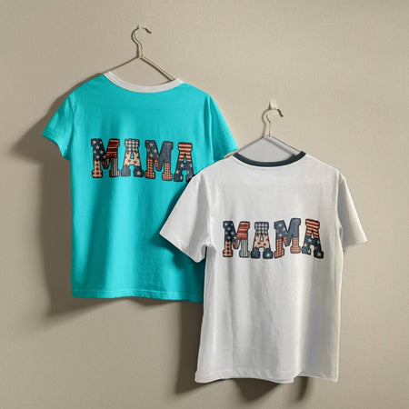 Two t-shirts hanging on a wall, one vibrant turquoise and one white, both printed with a 4th of July style red, white, and blue "MAMA" flag pattern graphic.