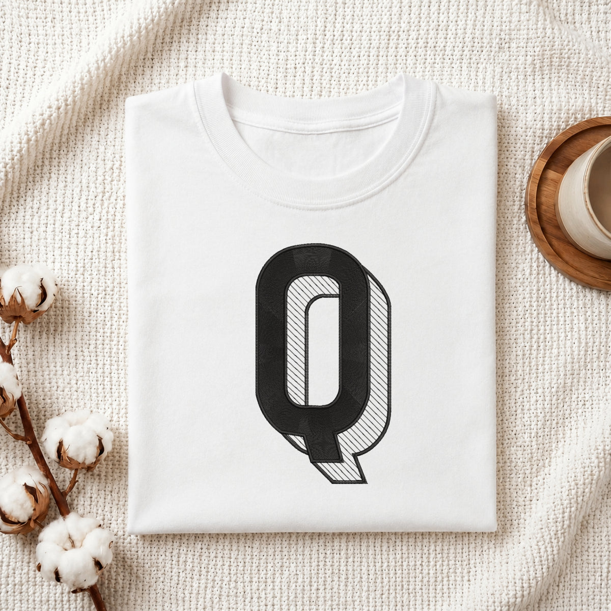 A folded white crewneck t-shirt with a large black graphic letter 'Q' on the front, styled on a textured cream knit blanket with cotton stems and a wooden cup in the background.
