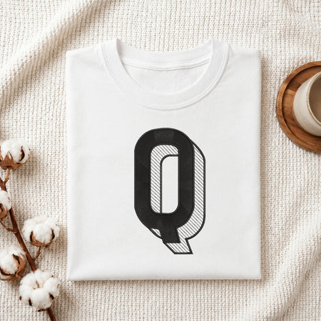 A folded white crewneck t-shirt with a large black graphic letter 'Q' on the front, styled on a textured cream knit blanket with cotton stems and a wooden cup in the background.