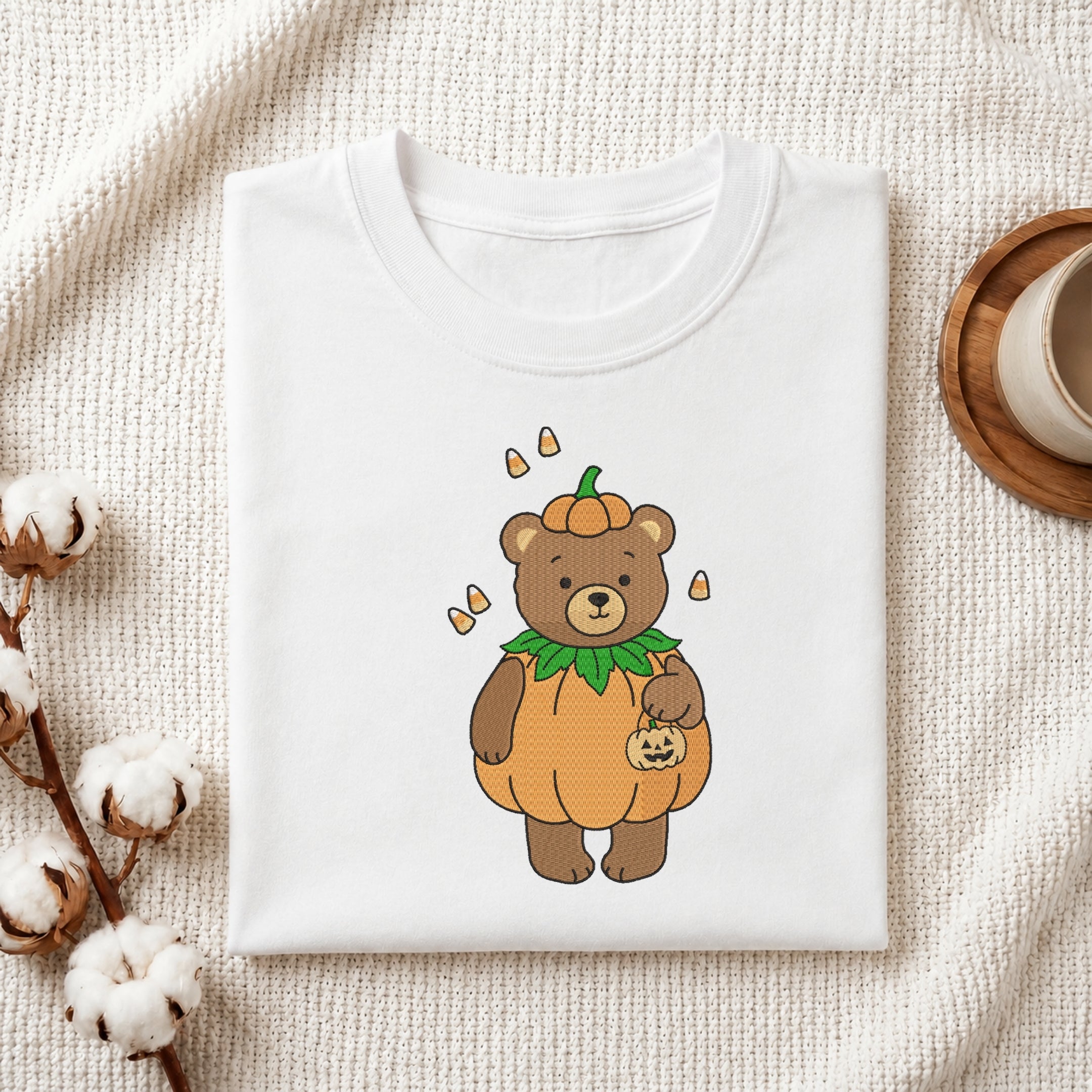 A neatly folded white crewneck t-shirt featuring a stitched Halloween bear design, styled with cotton branches and a wooden coaster.