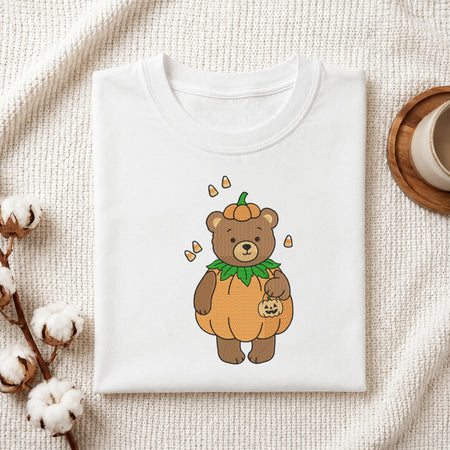 A neatly folded white crewneck t-shirt featuring a stitched Halloween bear design, styled with cotton branches and a wooden coaster.