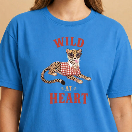 Smiling person wearing a white t-shirt featuring a trendy leopard wearing sunglasses, a gold chain, and a plaid shirt, blowing a pink bubblegum bubble, with the text "WILD AT HEART".