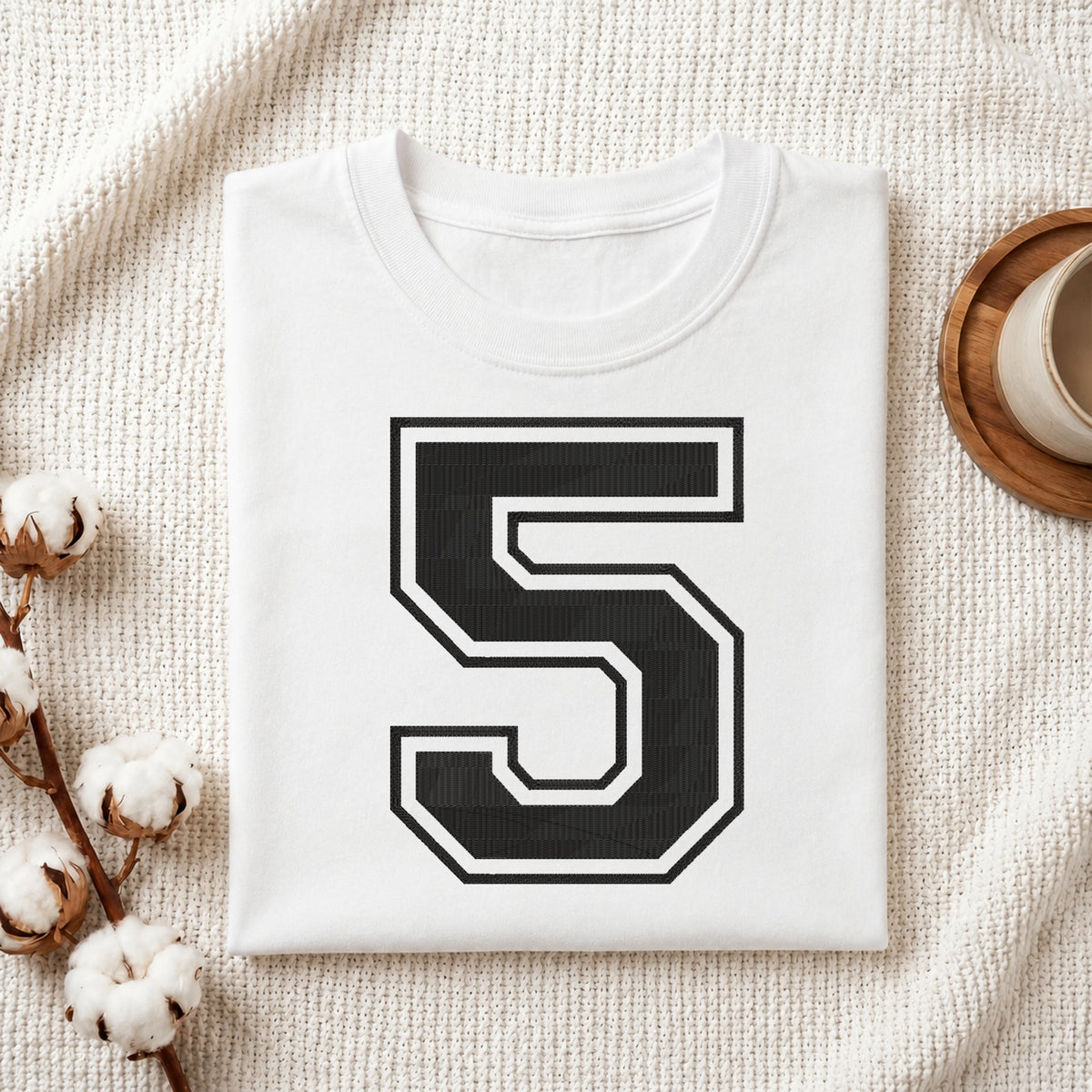 A folded white t-shirt with a large black and white number 5 printed on the front, styled on a textured cream blanket with cotton stems and a cup in the background.