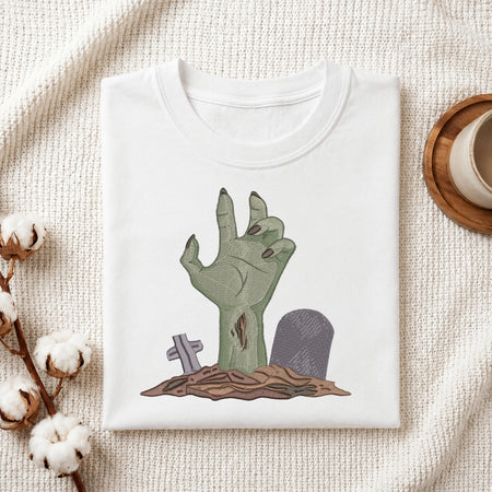 A folded white t-shirt with a zombie hand coming out of a grave illustration, styled on a textured cream fabric with cotton bolls and a cup on a wooden tray.