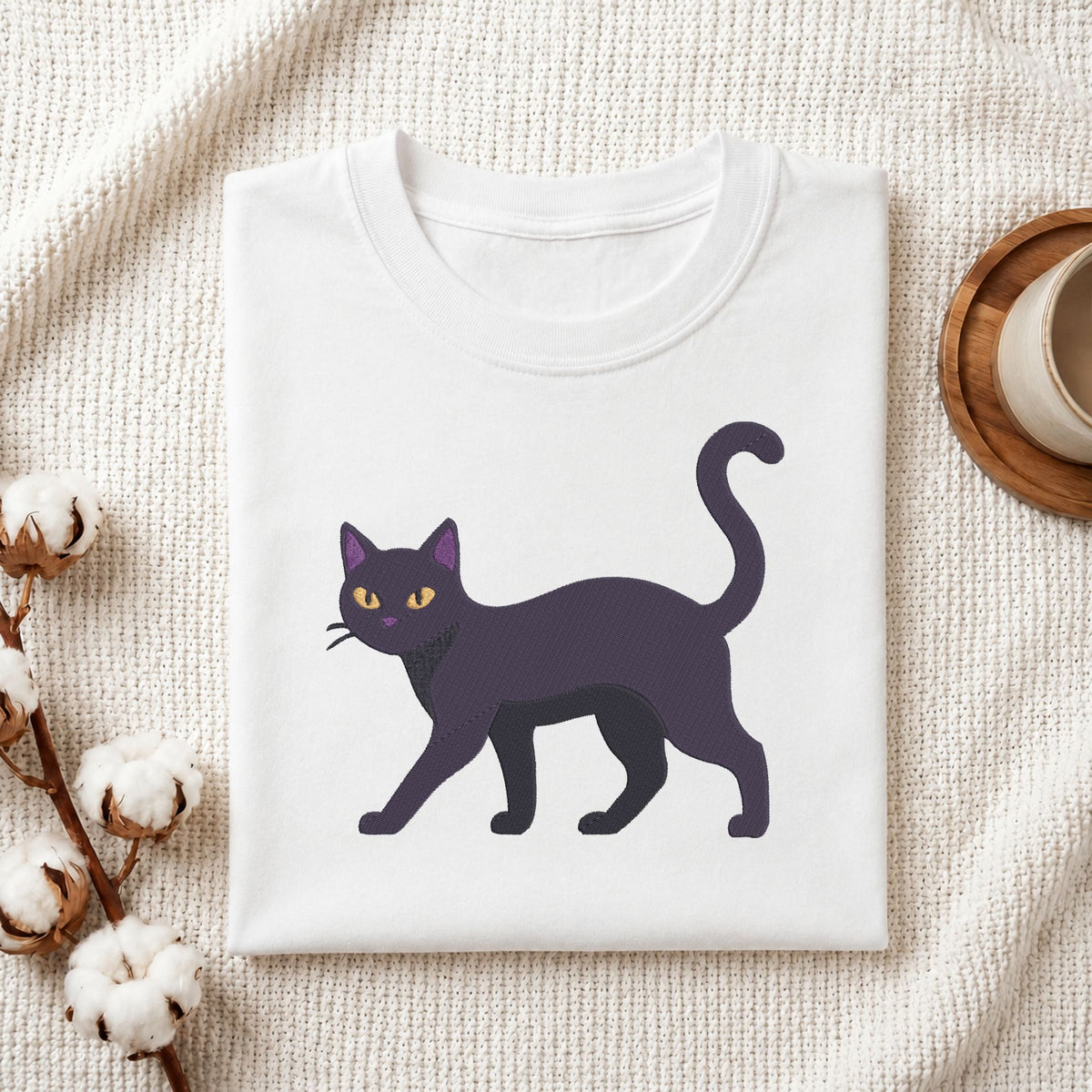 A folded white crewneck t-shirt featuring a graphic of a black cat with yellow eyes, styled on a textured cream fabric with cotton bolls and a wooden cup.