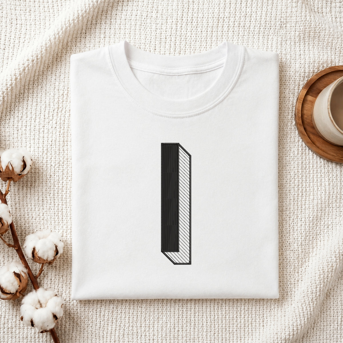 A folded white crewneck t-shirt with a minimalist black graphic of the number '1' styled on a textured cream fabric background with cotton bolls and a wooden cup.