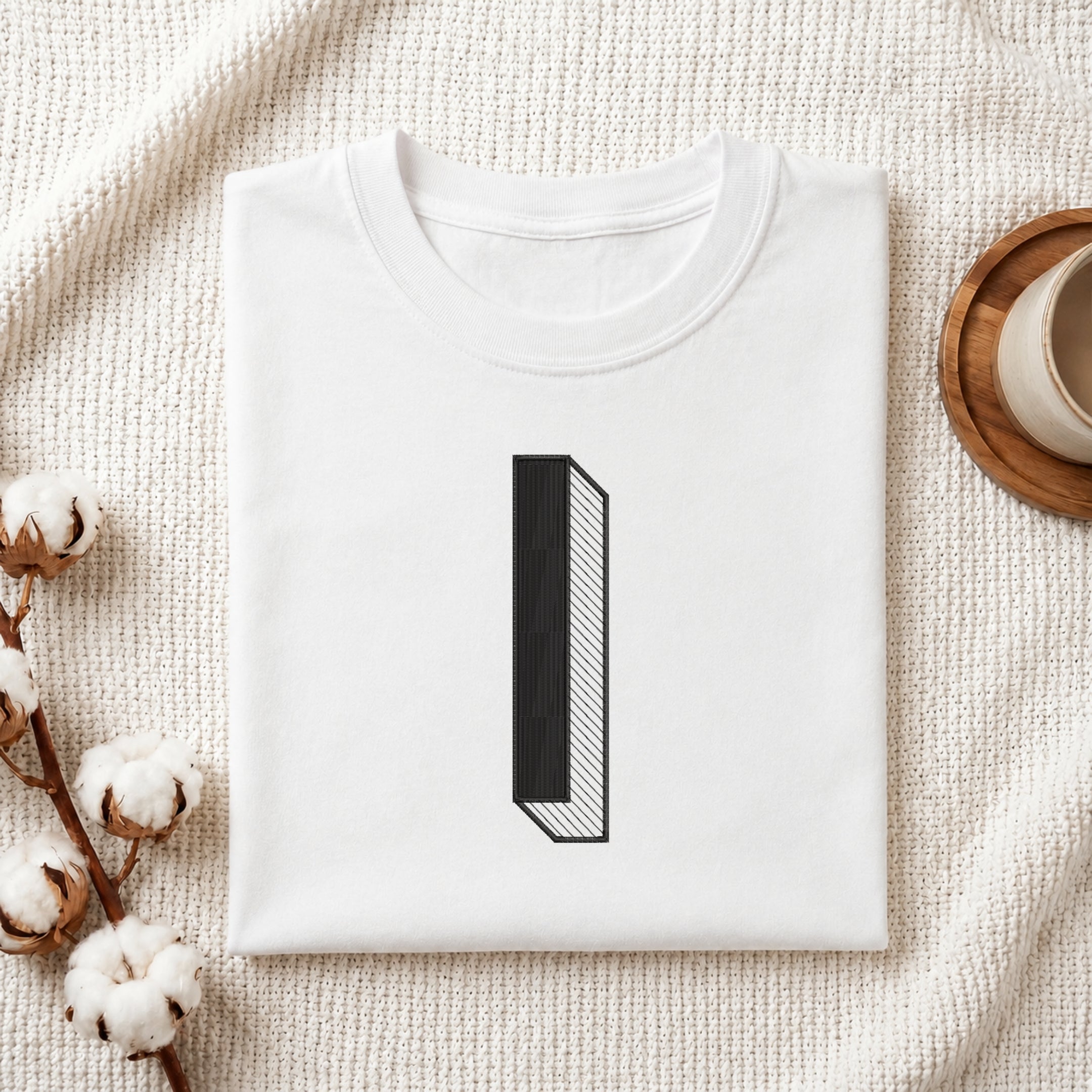 A folded white crewneck t-shirt with a minimalist black graphic of the number '1' styled on a textured cream fabric background with cotton bolls and a wooden cup.
