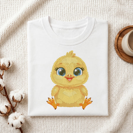 A folded white crewneck t-shirt featuring a cute cartoon illustration of a yellow baby chick with large blue eyes, styled on a textured cream fabric background with cotton bolls and a wooden cup.