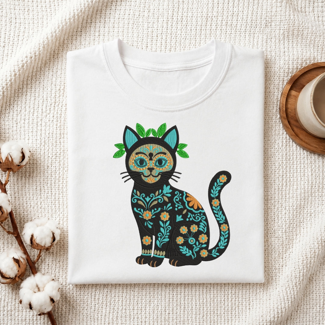 A folded white crewneck t-shirt featuring a colorful Dia de los Muertos cat graphic, styled on a textured cream knit blanket with cotton bolls and a wooden cup in the background.