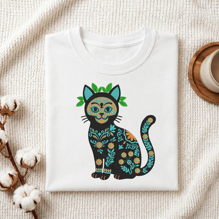 A folded white crewneck t-shirt featuring a colorful Dia de los Muertos cat graphic, styled on a textured cream knit blanket with cotton bolls and a wooden cup in the background.