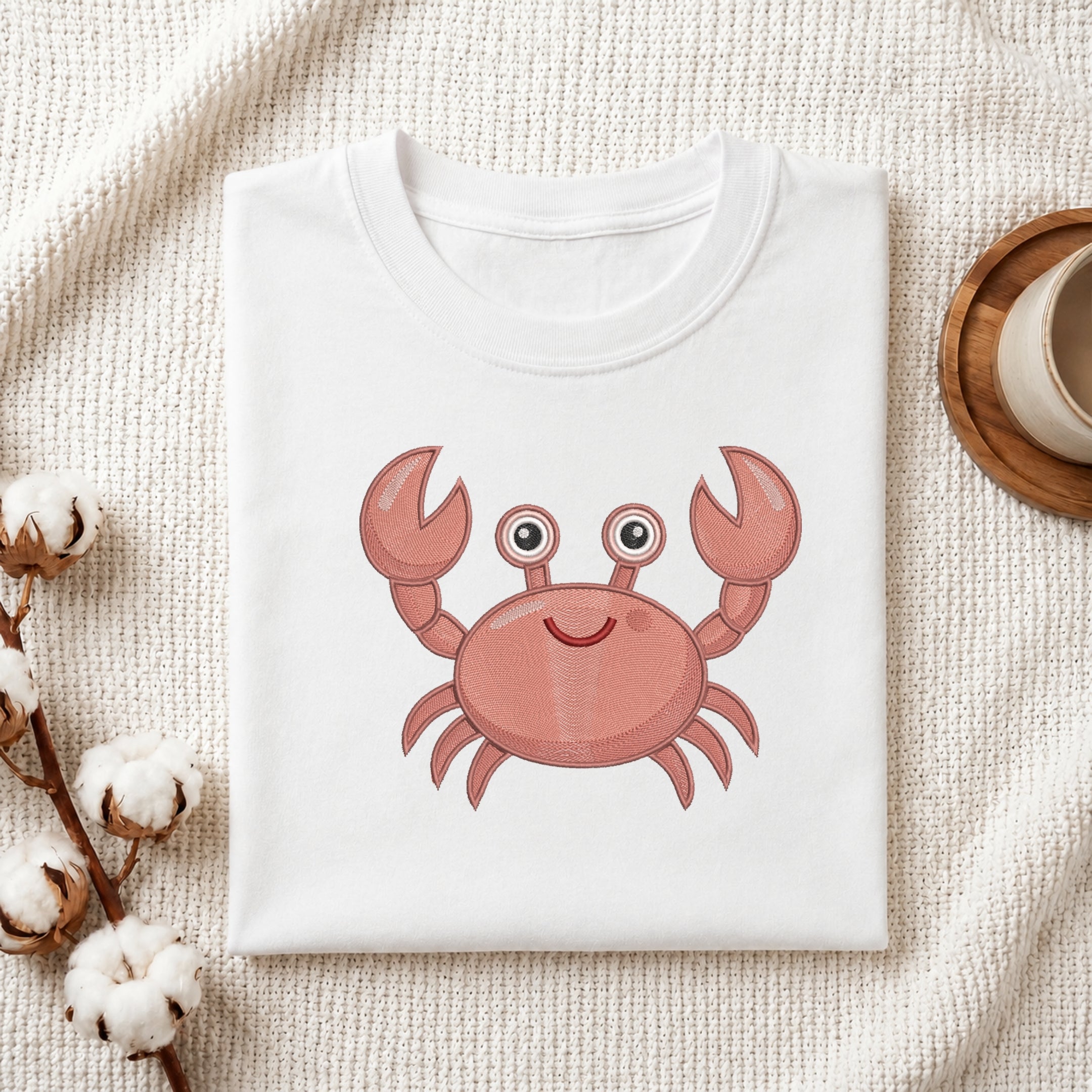 A folded white crewneck t-shirt featuring a cute pink cartoon crab graphic on the front, styled on a textured cream fabric with cotton bolls and a wooden cup.
