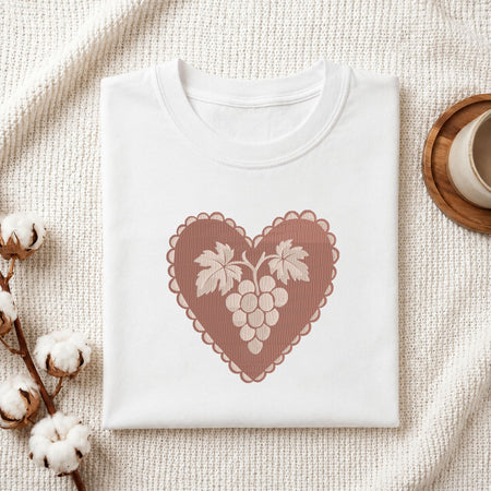 Folded white cotton t-shirt mockup featuring a heart-shaped grape cluster machine embroidery design, styled with cotton stems and a wooden cup.