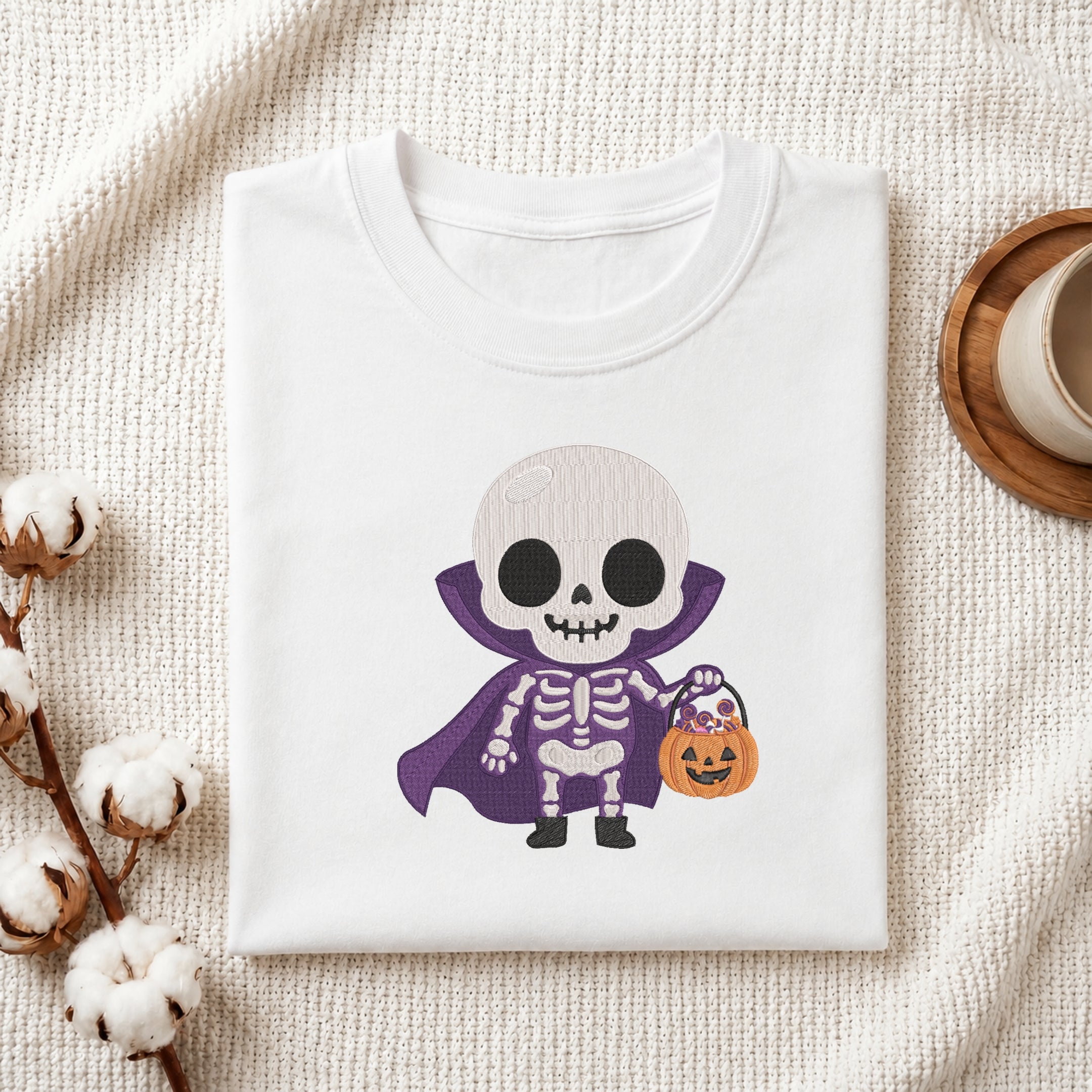 A folded white t-shirt with a cute cartoon skeleton Halloween graphic, styled on a textured cream fabric background with cotton bolls and a wooden cup.