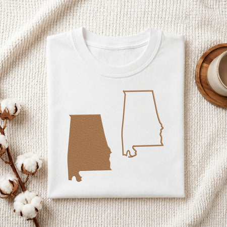 A folded white T-shirt with two brown Alabama state silhouette graphics on the front, styled on a textured cream fabric with cotton bolls and a cup of coffee nearby.