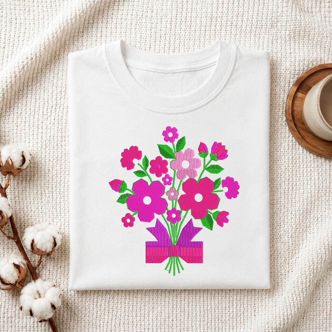 A folded white crewneck t-shirt with a colorful graphic of a flower bouquet in pink, purple, and green, styled on a textured cream fabric with cotton bolls and a wooden cup.