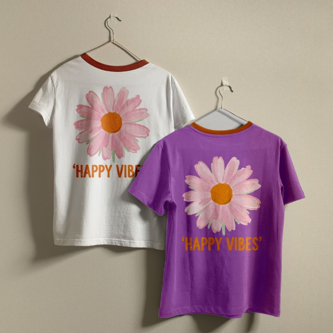 Two ringer t-shirts hanging on hangers against a plain beige wall, showing the front and back designs. The left shirt is white with a red collar and features a pink daisy graphic with the text 'HAPPY VIBE' in orange.