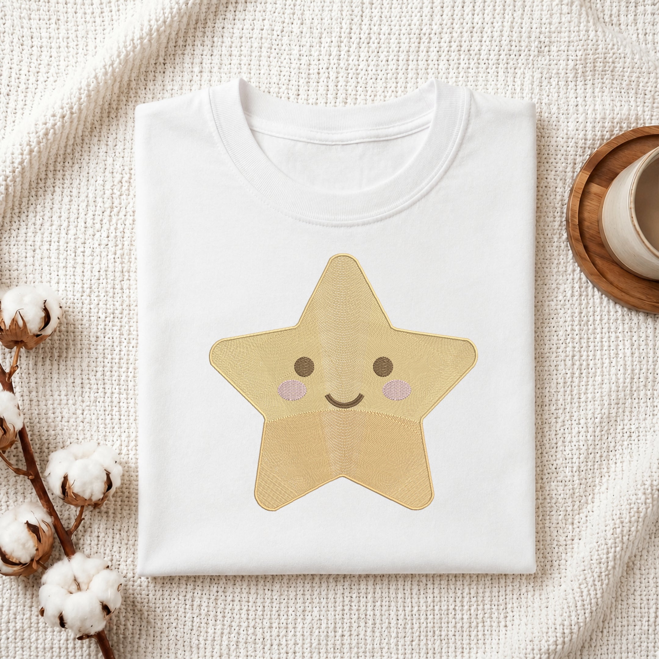 A folded white crewneck T-shirt with a cute embroidered star graphic in shades of yellow, beige, and pink, styled on a textured cream knit blanket with cotton bolls and a wooden cup in the background.