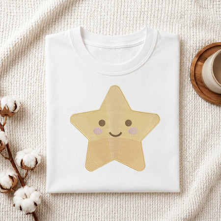 A folded white crewneck T-shirt with a cute embroidered star graphic in shades of yellow, beige, and pink, styled on a textured cream knit blanket with cotton bolls and a wooden cup in the background.