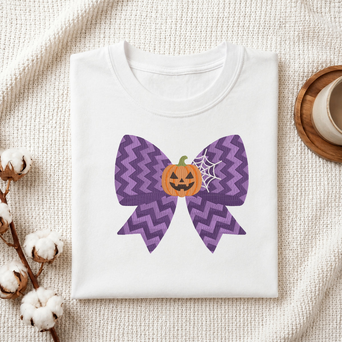 A neatly folded white t-shirt featuring the embroidered purple chevron bow, jack-o'-lantern, and spiderweb design, displayed on a textured white knit background with cotton stems and a wooden cup.
