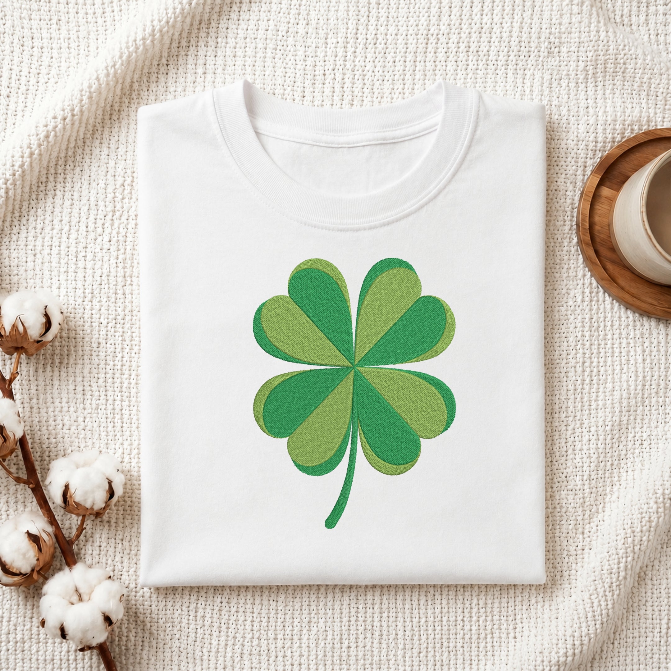 A folded white crewneck T-shirt with a green shamrock graphic printed on the chest, styled on a textured cream fabric with cotton bolls and a wooden cup in the background.