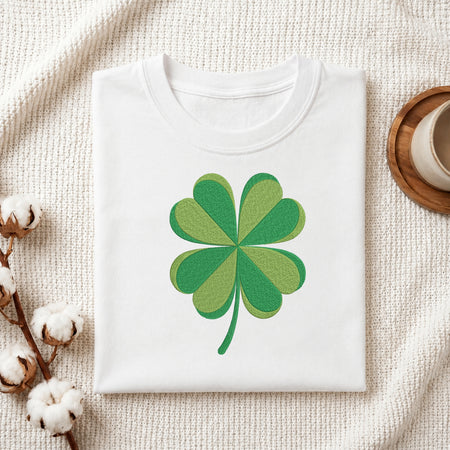 A folded white crewneck T-shirt with a green shamrock graphic printed on the chest, styled on a textured cream fabric with cotton bolls and a wooden cup in the background.