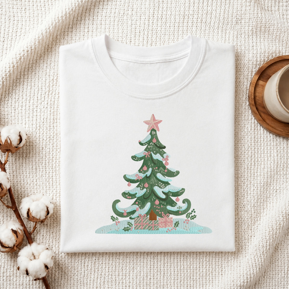 A folded white crewneck t-shirt with a colorful Christmas tree graphic, styled on a textured cream knit blanket with cotton bolls and a wooden mug in the background.