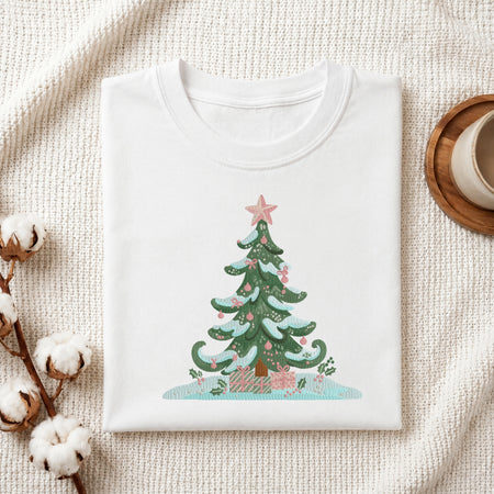 A folded white crewneck t-shirt with a colorful Christmas tree graphic, styled on a textured cream knit blanket with cotton bolls and a wooden mug in the background.