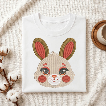 A folded white crewneck sweatshirt or t-shirt featuring a large embroidered bunny face graphic on the front, styled on a textured cream fabric with cotton stems and a wooden cup in the background.