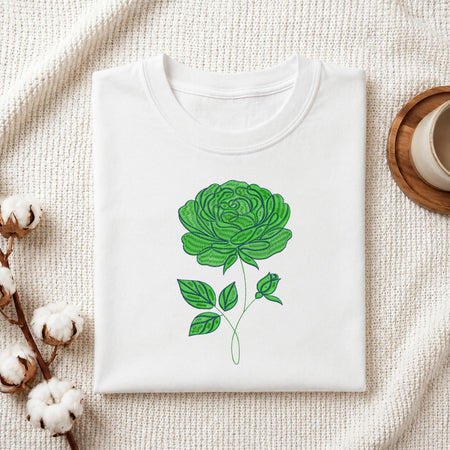A folded white crewneck t-shirt featuring a green rose graphic on the front, styled on a textured cream fabric with cotton bolls and a wooden cup in the background.